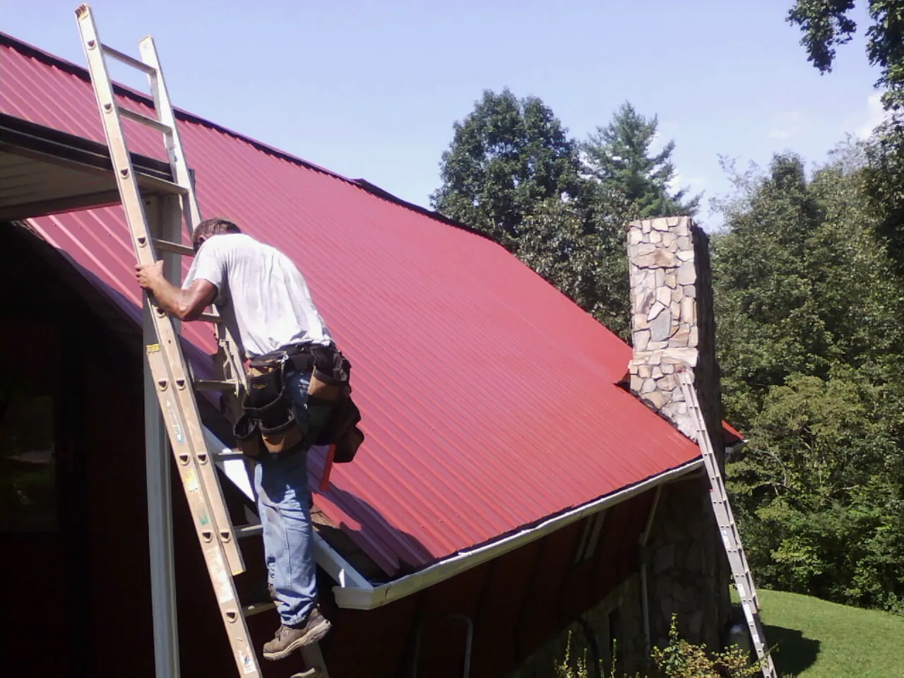Roofer on a ladder beside a red metal roof and stone chimney — Soffit & Fascia Repair by Roofing Brothers in Thorn Hill